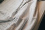 Roupa de cama em algodão branco com luz suave da manhã, ilustrando a escolha entre percal e cetim