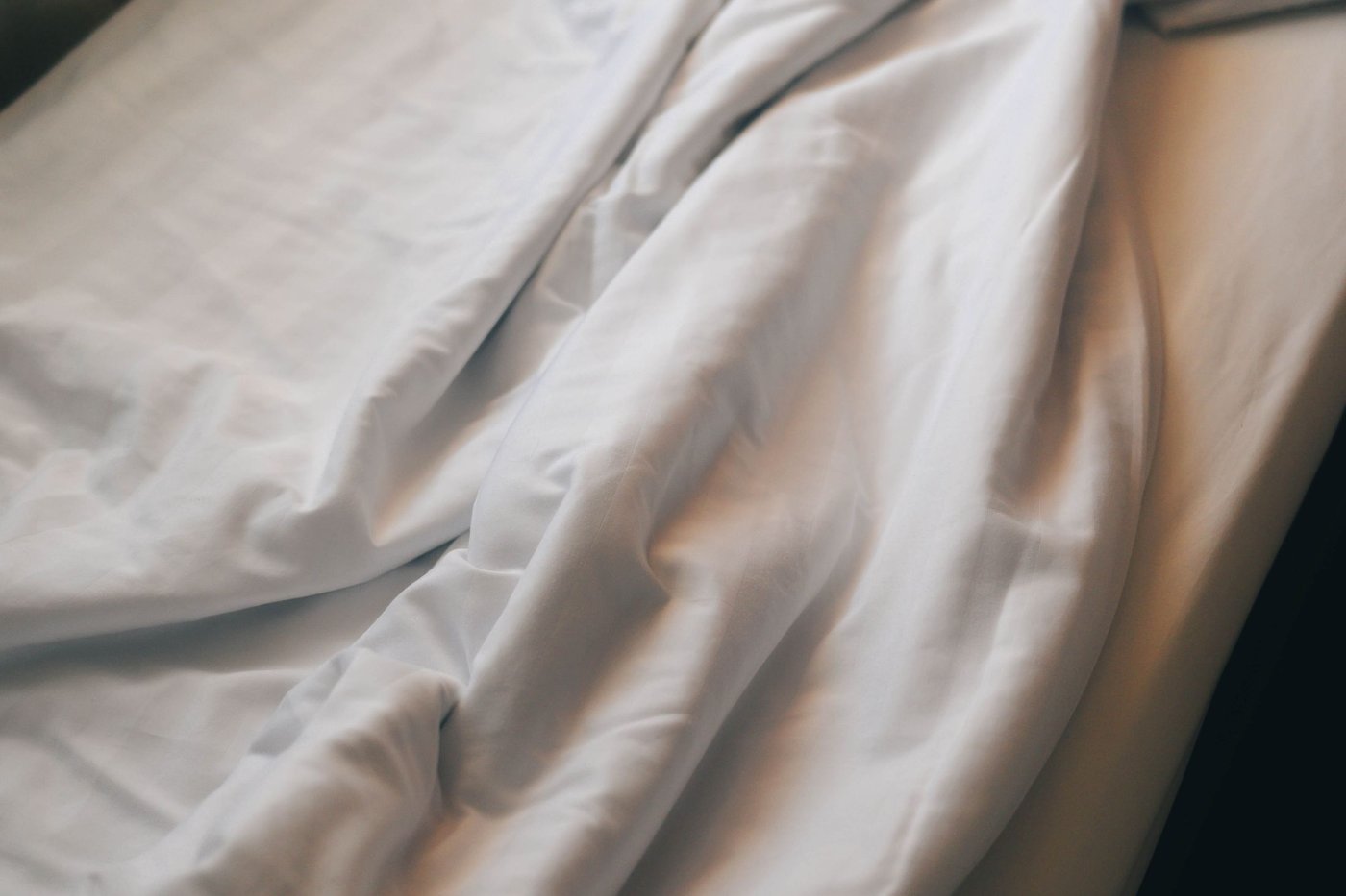 Roupa de cama em algodão branco com luz suave da manhã, ilustrando a escolha entre percal e cetim